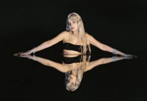 kim petras leaning over a mirror with arms outstretched wearing a black bandeau top