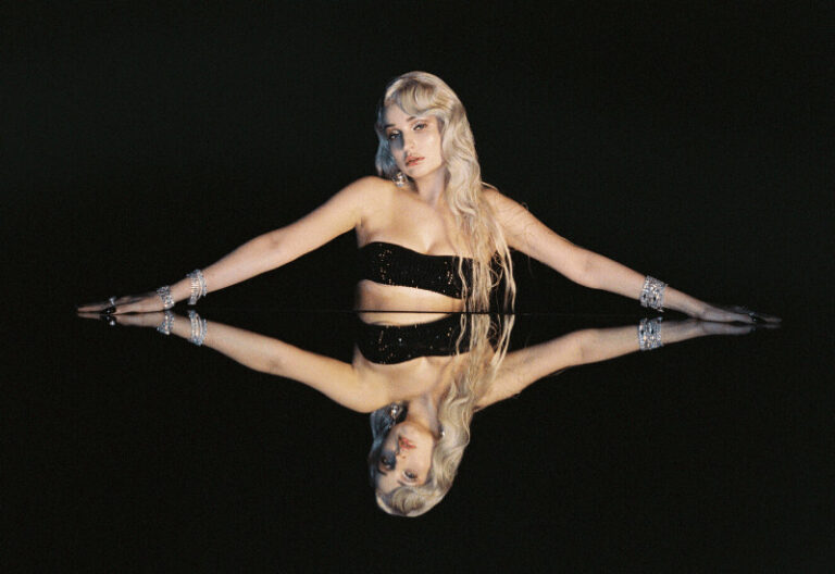kim petras leaning over a mirror with arms outstretched wearing a black bandeau top