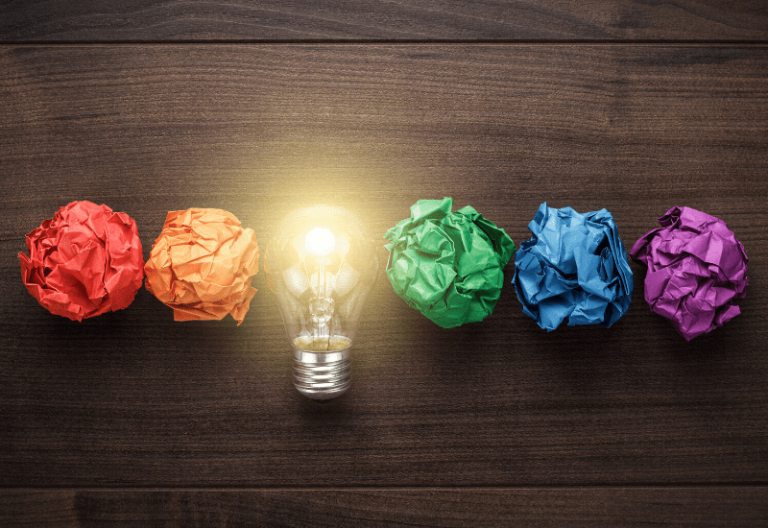 red orange green blue purple crumpled paper with a yellow light bulb in the middle on a brown faux wood background