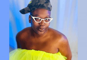 TITUSS BURGESS in a yellow top, camouflage baseball cap and white glasses against a blue background