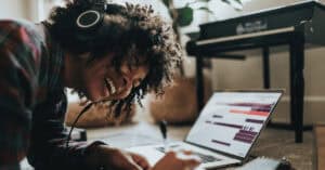 Black woman with afro enjoys music through earphones while laying in front of a laptop
