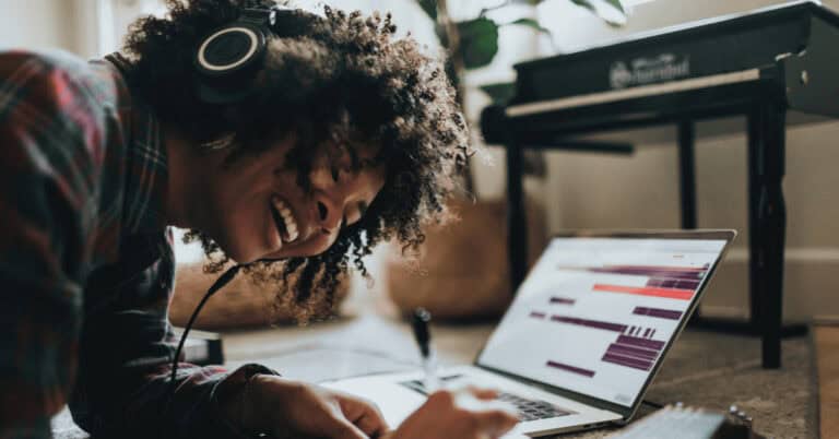 Black woman with afro enjoys music through earphones while laying in front of a laptop