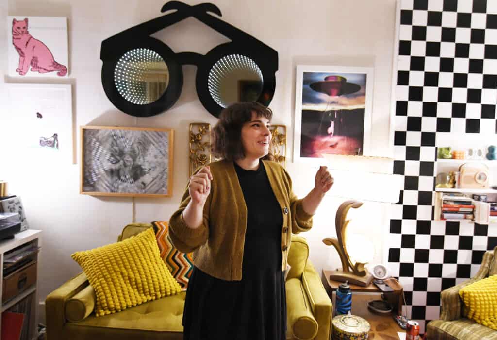 Sara Moseley stands smiling in colorful room with checkered wall and many knick knacks behind her.