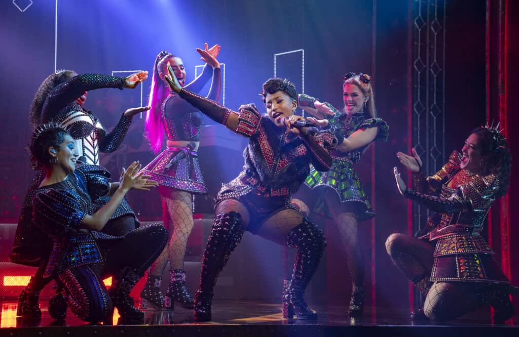 El elenco de The Six se forma mientras canta en un escenario iluminado en rojo.