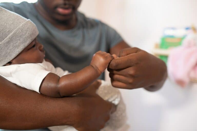 Un hombre negro con camisa blanca sostiene la mano del bebé.