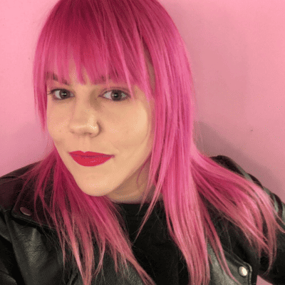 Kelly Seagraves in front of a pink background with pink hair, pink lipstick and a black leather jacket.