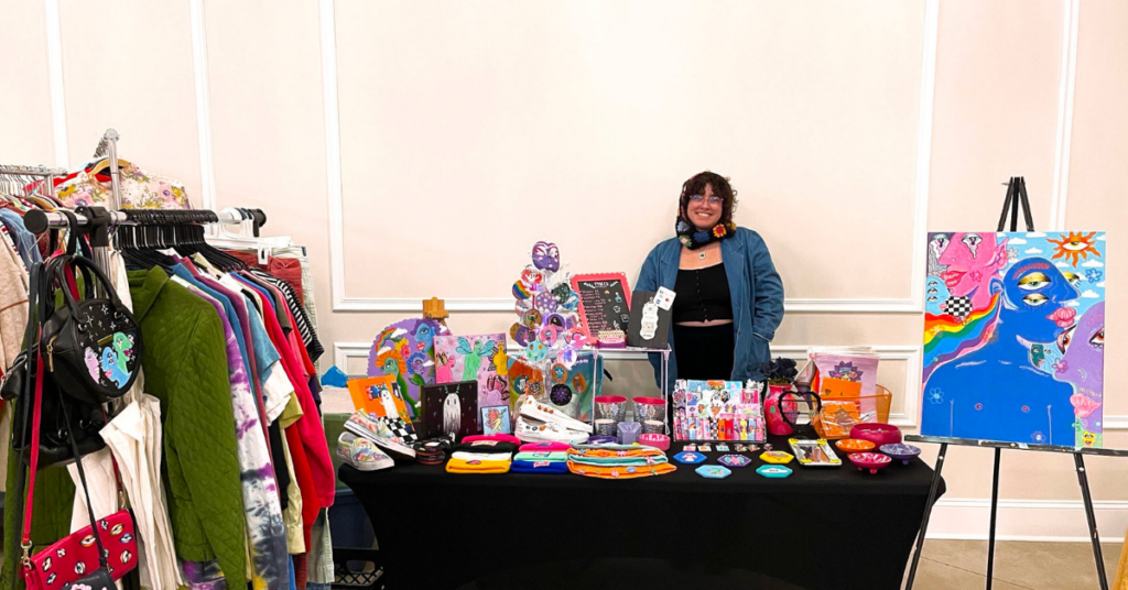 Shira Mae stands behind a booth covered in their original artwork and designs and clothes for sale at a market. They stand in front of a cream colored wall