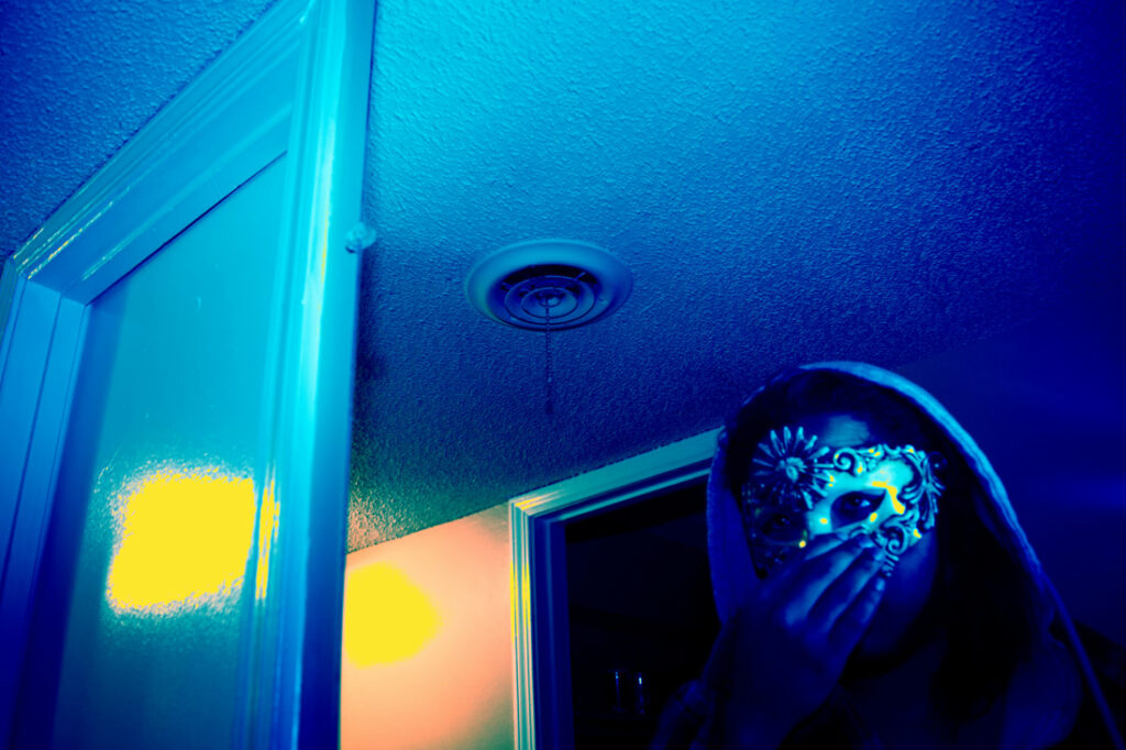 Lo Norman masked, self portrait in blue light with their hand covering their face.