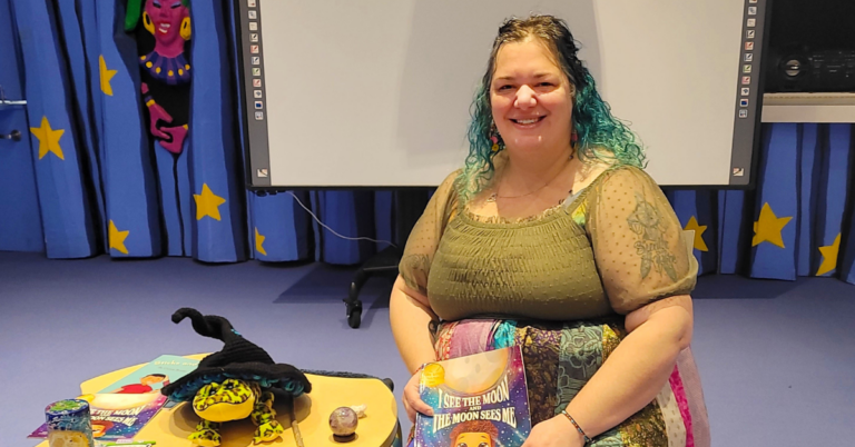Cassie Brooks, a White woman with long hair sits in front of large screen and blue background holds book by herself. Sits beside yellow table with knick knacks on it