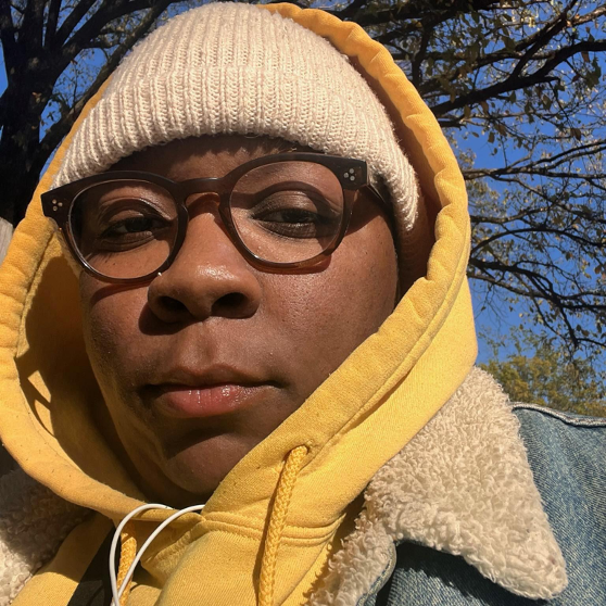 Black trans person in yellow hoodie with brownish glasses