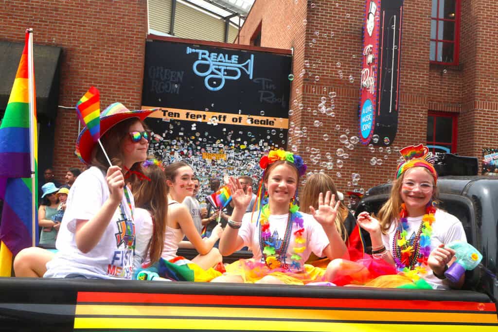 Performers in rainbow accessories and colorful costumes wave from a parade float. — focuslgbt.com