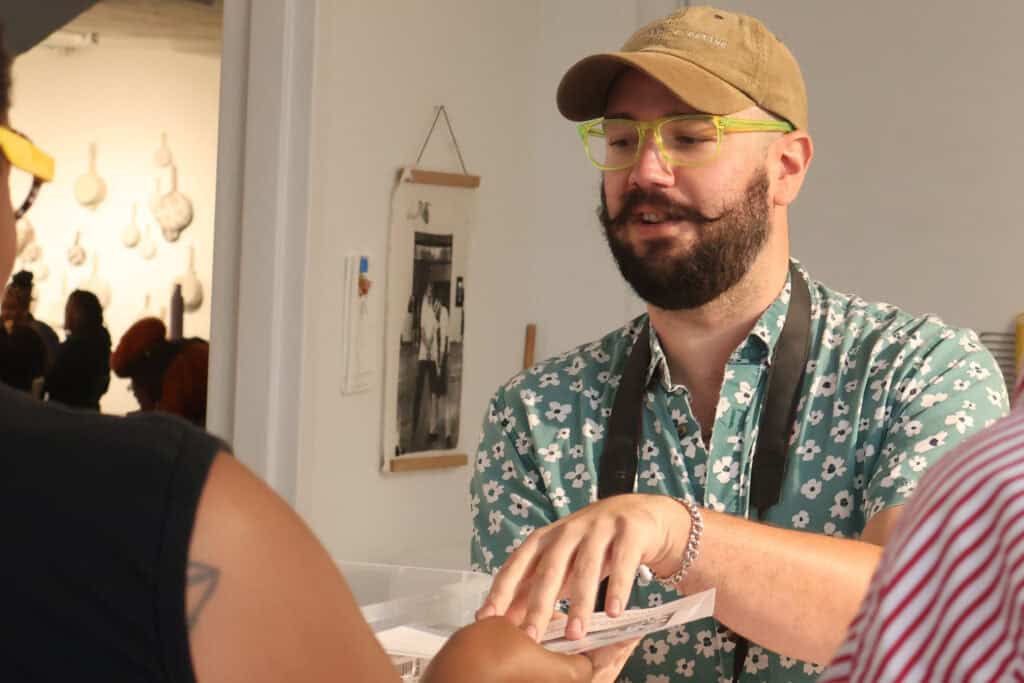 Man with glasses, beard, and tan cap hands a card to a person in black at an indoor event. — focuslgbt.com